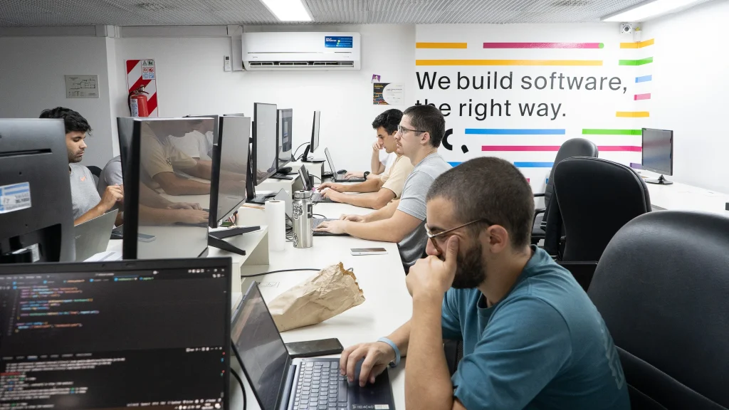 A wide-angle, modern office interior shot showing a bright, open workspace where several Crombie employees are working on computers. This environment supports high-focus AI software development and efficient delivery of AI-powered solutions for retail and ecommerce.
